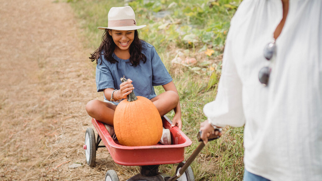 A young girl riding in a red wagon holding a big orange pumpkin