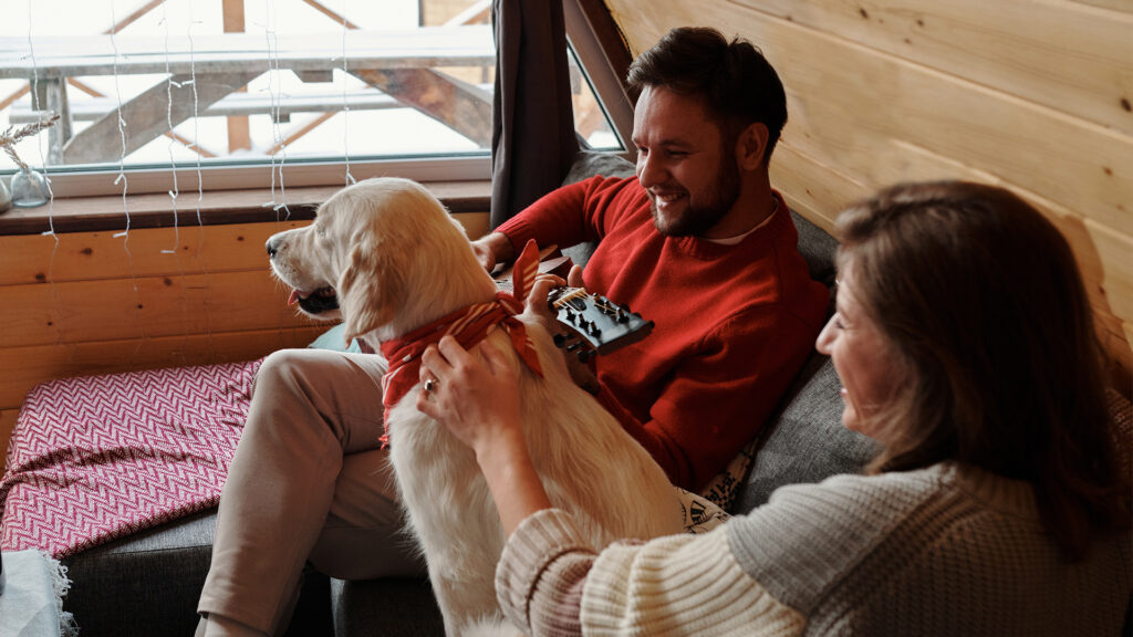 A man in a red sweater sitting on the couch in a cabin with a guitar in hand beside a white dog with a red bandana and a woman