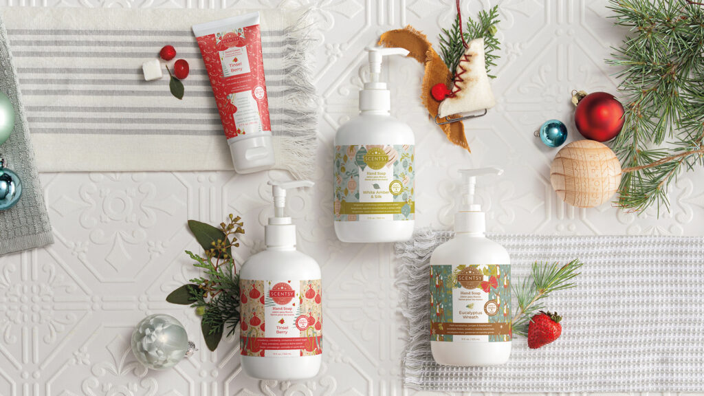 Scentsy holiday collection body products including Holiday Hand Soap 3-pack, featuring Eucalyptus Wreath, Tinsel Berry and White Amber & Silk fragrance and the Tinsel Berry Hand Cream laid out on a white back drop with christmas ornaments and pine decor