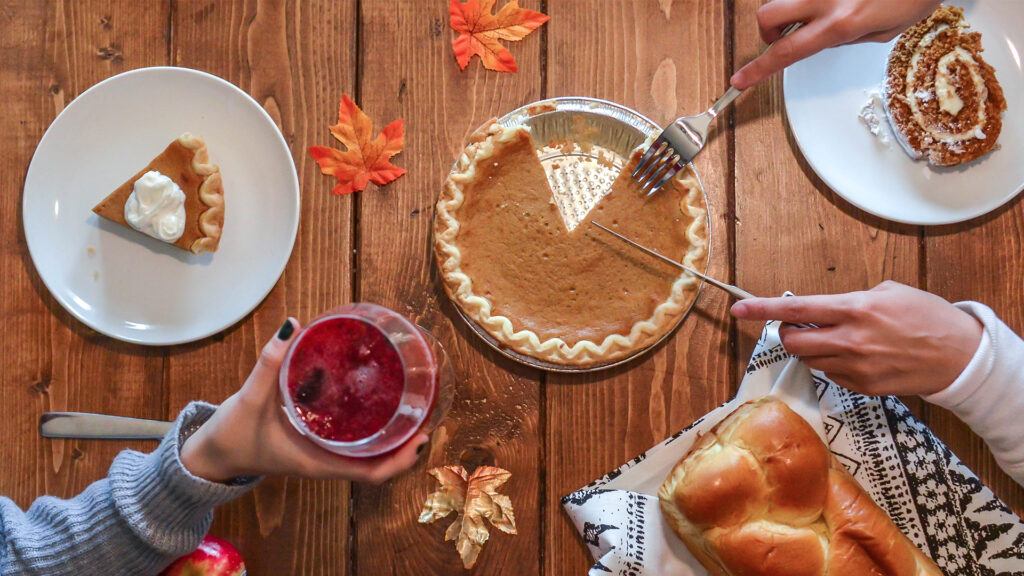 Overhead shot of two people arms over the dinner table cutting up a pumpkin pie beside a loaf of bread and two plates one with a pumpkin roll and one with a slice of pumpkin pie holding a red beverage in her hand.
