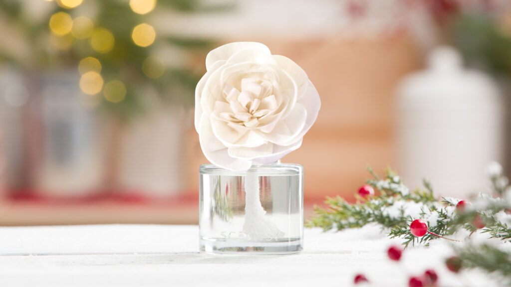 scentsy fragrance flower sitting on a white table beside pine and red berry foliage with a christmas tree blurred in the background
