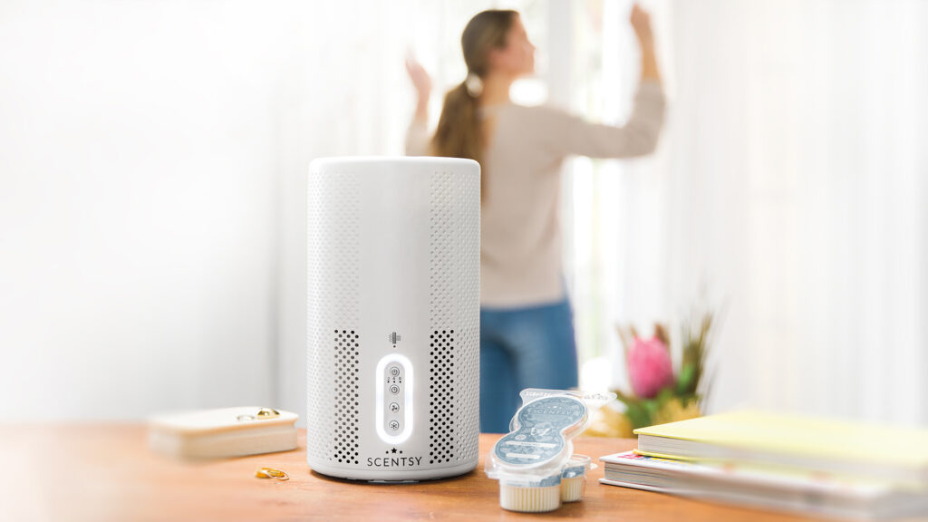 A scentsy air purifier and scent pods sitting on a desk beside notebooks with a woman in the background opening her curtains to let in sunlight