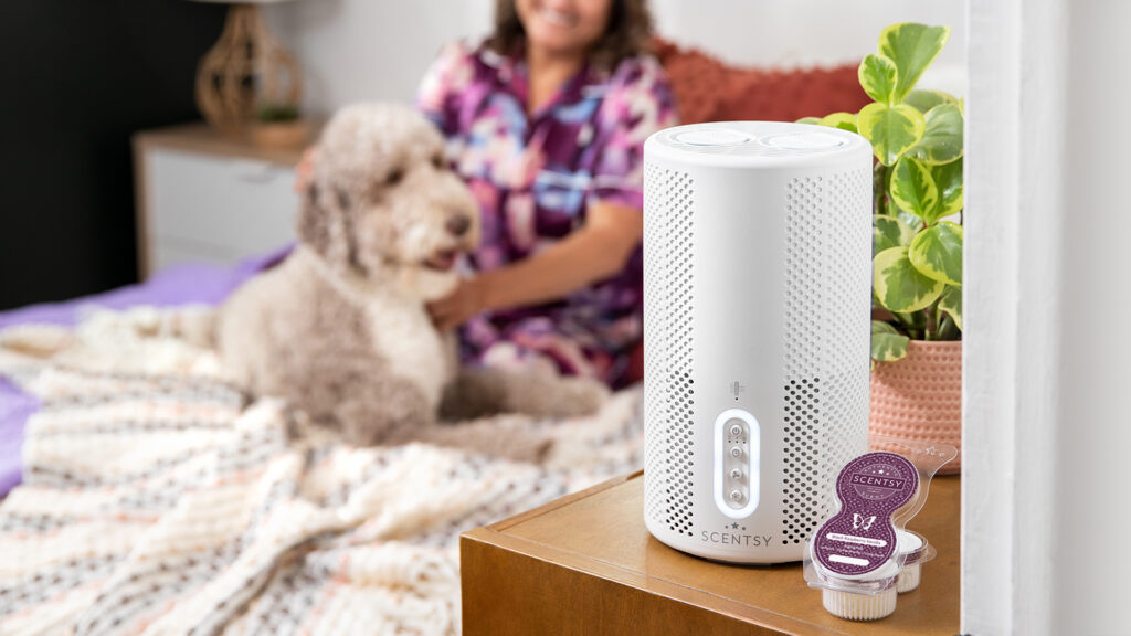 Scentsy air purifier sitting on a bedroom dresser beside scentsy pods and a house plant in a pink pot with a woman and her doodle dog sitting in the background on a bed
