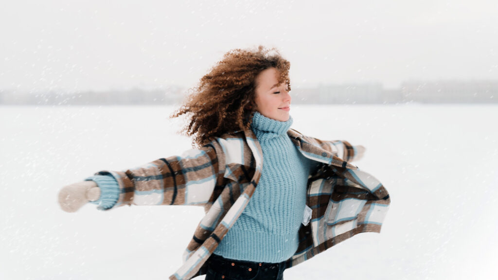 A woman in a baby blue turtleneck sweater, black and brown plaid jacket and white mittens with her arms out embracing the snow while standing in a field filled