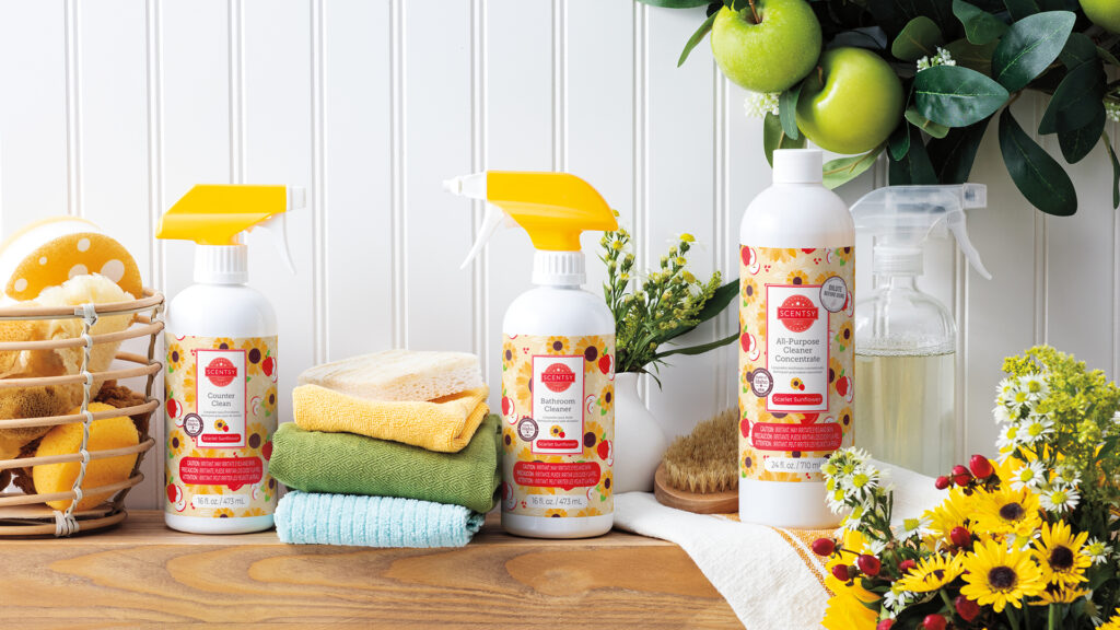Scentsy cleaning products sitting on a bathroom counter in scarlet sunflower fragrance with sponges, hand towels, flowers and a green leafy wreath with apples.