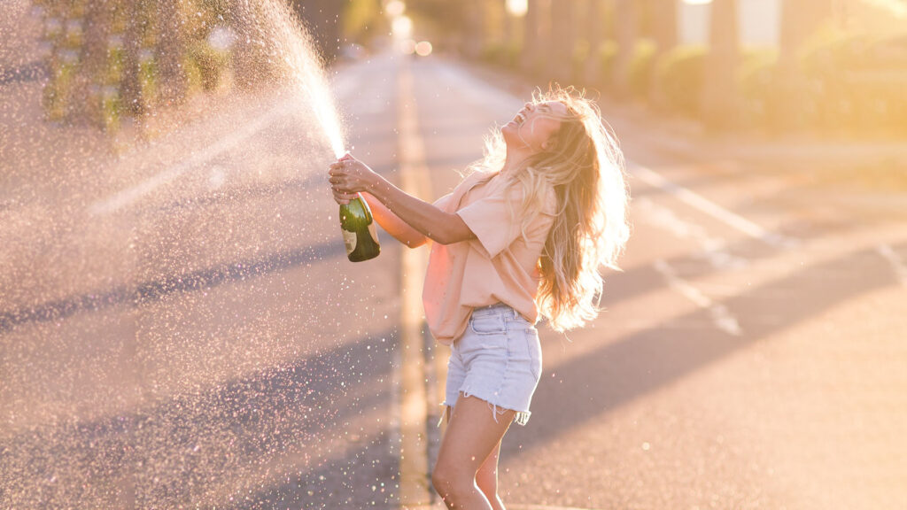 A girl standing in the middle of the street on a sunny day in shorts and a tshirt shaking and spraying a bottle of champagne into the air 