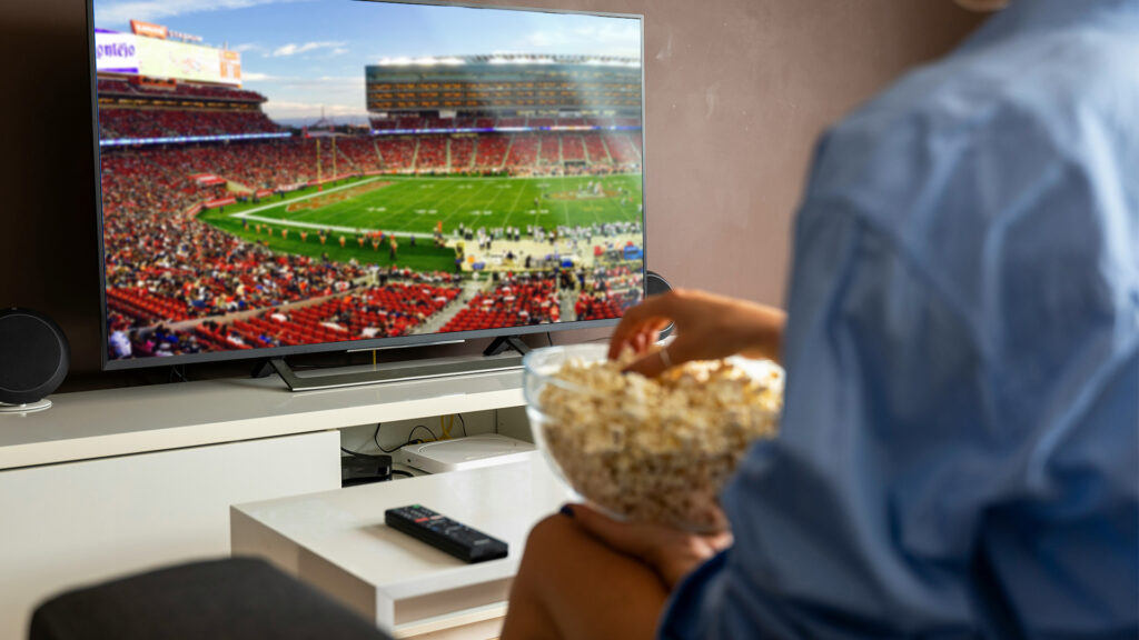 A woman sitting on a couch with a bowl of popcorn in her lap watching a football game