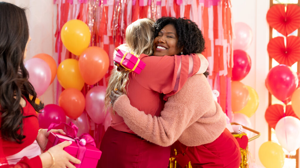 Two women exchanging gifts and hugging in front of a festive valentines day backdrop at a valentines day party