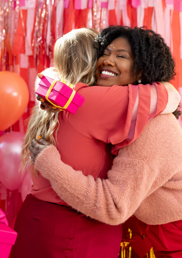 Two women exchanging gifts and hugging in front of a festive valentines day backdrop at a valentines day party