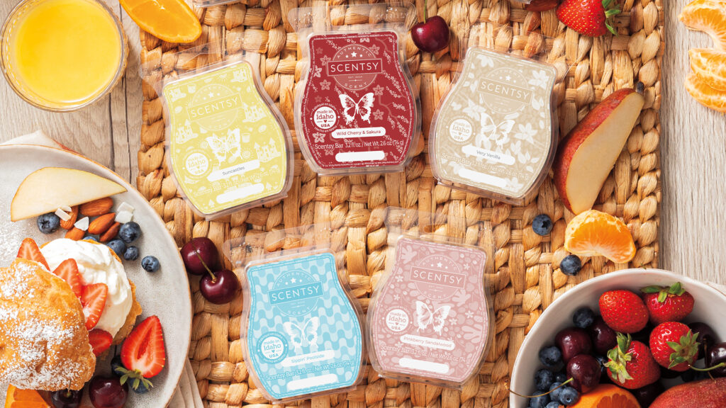 five scentsy wax melts in different fragrances sitting on a wicker placemat beside breakfast spread with orange juice, bowl of fruit and whipped cream on a breakfast biscuit