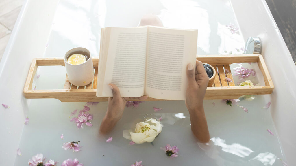 A woman laying in the bath filled with flowers and bath soap reading a book over a a wooden bath table on it is a cup of lemon tea and bowl of blueberries 