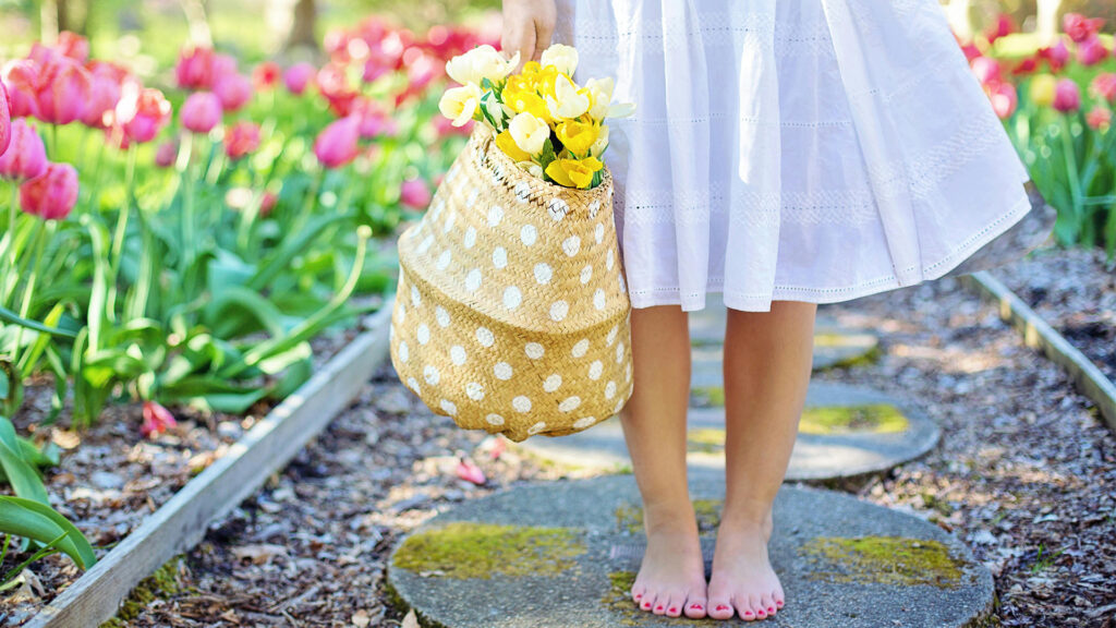 a woman standing in a tulip garden on a circle cement paver holding a wicker white polka dotted basket with yellow tulips in a white flowy summer dress