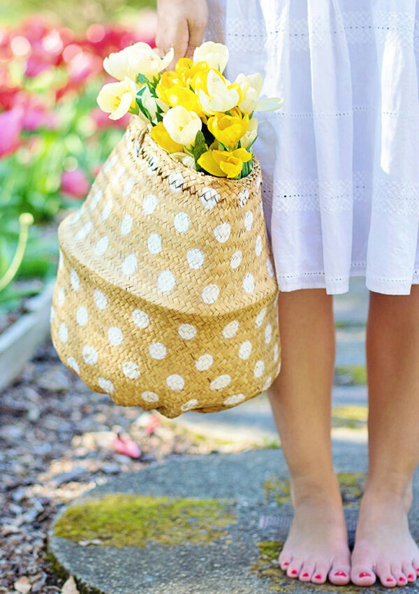 a woman standing in a tulip garden on a circle cement paver holding a wicker white polka dotted basket with yellow tulips in a white flowy summer dress
