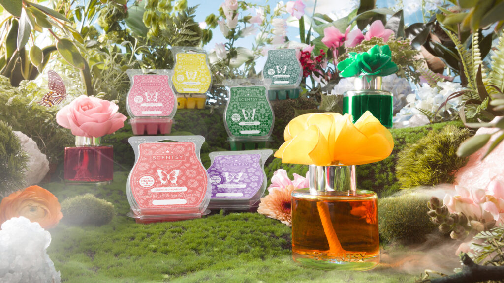Three Fragrance Flowers and six Scentsy Wax Bars inspired by Blossoms of Fragrance, as well as flowers and gardens around the world sitting in a whimsical setting with butterflies, moss, ferns, peonies and more blossoming flowers lit up by sunshine