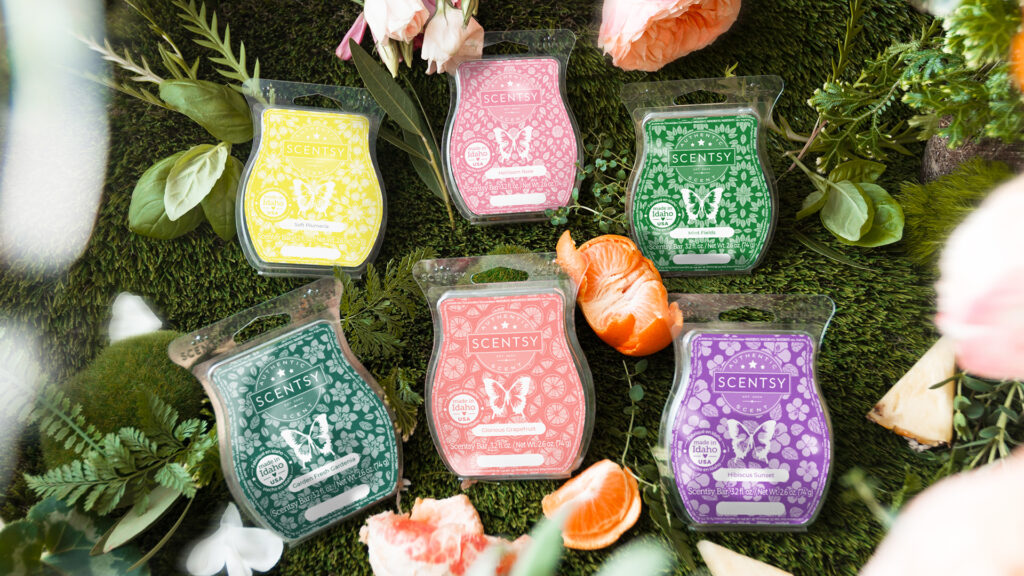 The six wax bars in Scentsy's Happiness Blooms Collection including Garden Fresh Gardenia, Glorious Grapefruit, Heirloom Rose, Hibiscus Sunset, Mint Fields and Soft Plumeria sitting on a mossy floral background resembling a whimsical forest