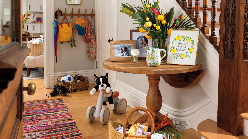 Entry way of a home with a wooden table with a big bright flower bouquet with lemons, picture frames, fragrance flower, lemon pitcher wax warmer, basket of towels and hand cream, kids tricycle with a scentsy buddy sitting on it and coat rack with a hat, coats, backpack buddy clip, and umbrella
