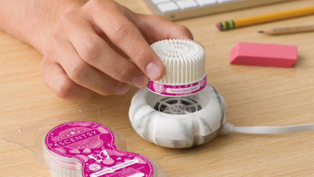 A hand dropping a summer berry melon scented scentsy pod into a mini fan diffuser on an office desk beside a keyboard and pencils
