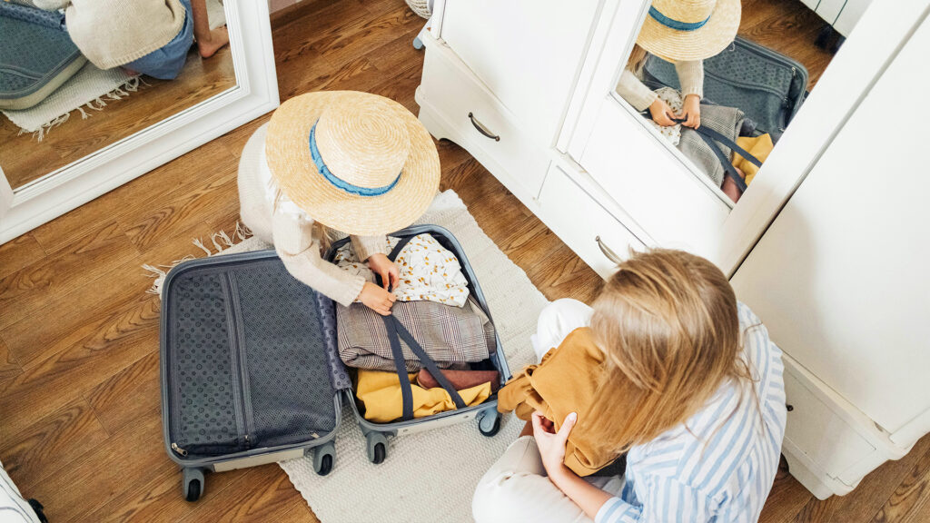 A woman and her daughter wearing a straw beach hat sitting on a bedroom floor packing a suitcase 