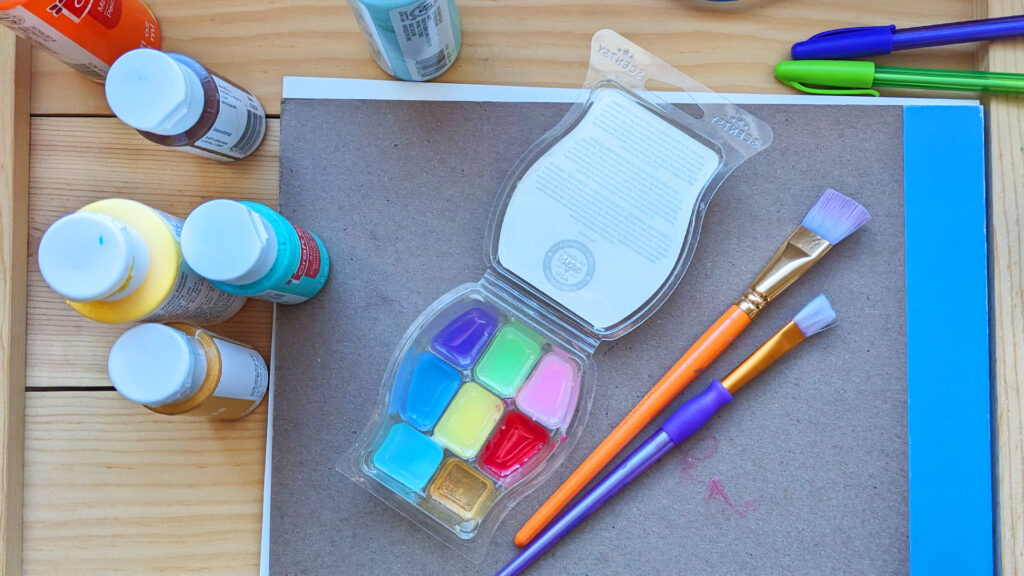 A repurposed scentsy wax bar container being used as a paint tray with different colors in each square beside paint containers and paint brushes