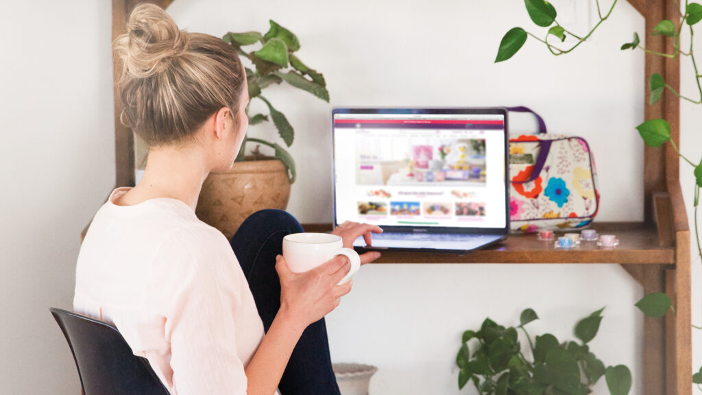 A woman holding a cup of coffee sitting at her desk scrolling on her computer on scentsy.com