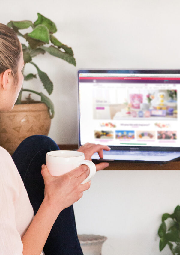 A woman holding a cup of coffee sitting at her desk scrolling on her computer on scentsy.com