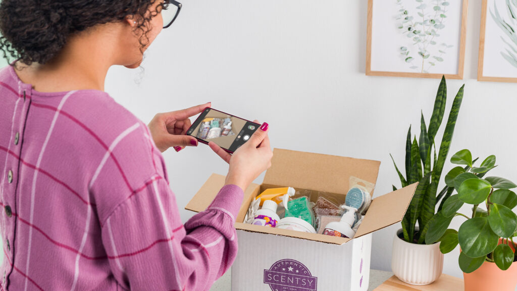 A woman taking a photo of her scentsy whiff box with green plants and pictures  of green plants in the background