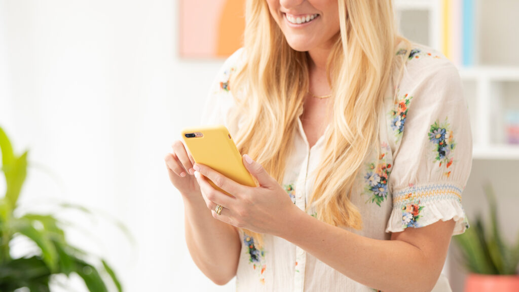 A blonde woman holding a phone with a yellow case smiling while scrolling
