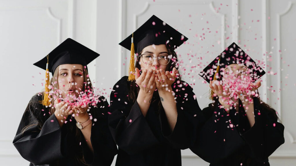 three girls dressed in caps and gowns blowing pink white and red confetti out of their hands