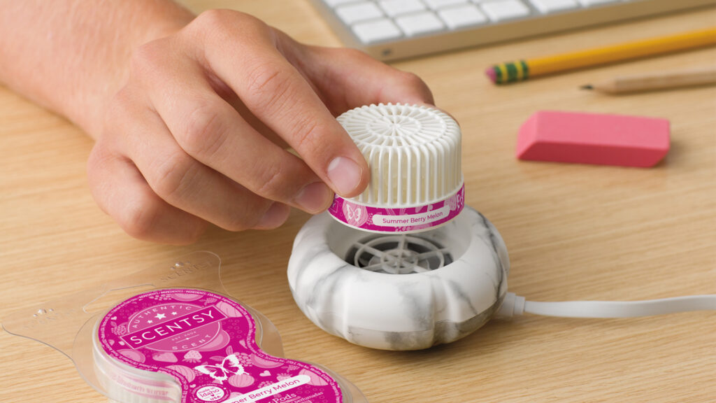 A hand dropping a summer berry melon scented scentsy pod into a mini fan diffuser on an office desk beside a keyboard and pencils