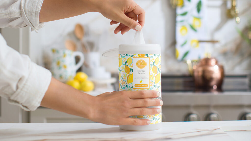 A woman pulling a cleaning wipe out of the scentsy swipes container in squeeze the day fragrance sitting on a kitchen counter with cooking utensils. lemon pitcher, a stove, gold kettle and lemons in the background