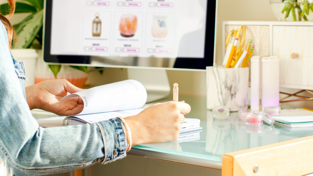 Someone sitting at a computer desk with Scentsy fragrance testers, taking notes in a fragrance journal and referencing the Scentsy website.