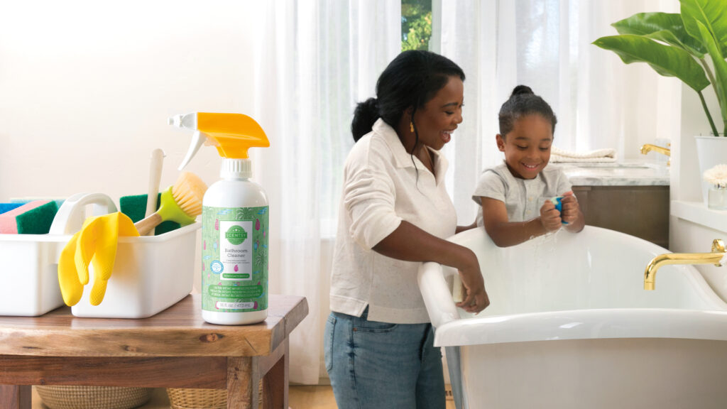 Scentsy clean and laundry products sit on a side table as a Mother teaches her child to clean a bath tub.