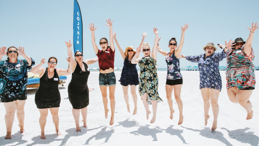 A group of women on the beach jump in the air, hands held up in excitment.