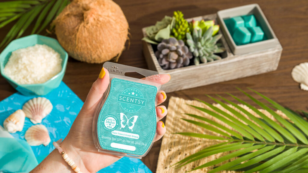 A hand holding the Scentsy Palm Trees and Ocean Breeze wax bar with a Suc-cute-lent Wax Warmer displayed on a beach themed table.