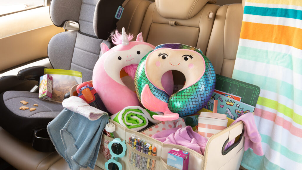 A crate of road trip accessories sits in the back seat, overflowing with cups towels, and Unicorn and mermaid Scentsy Buddy Travel Pillows.