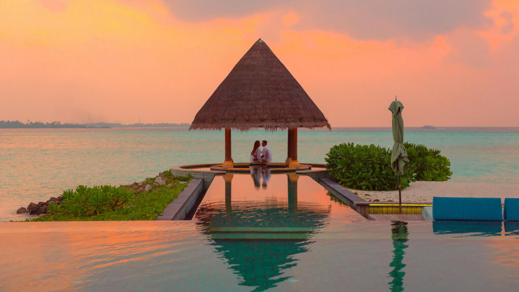 A couple sitting in a romantic, ocean-side gazebo watching the sunset together, water reflecting the orange clouds.