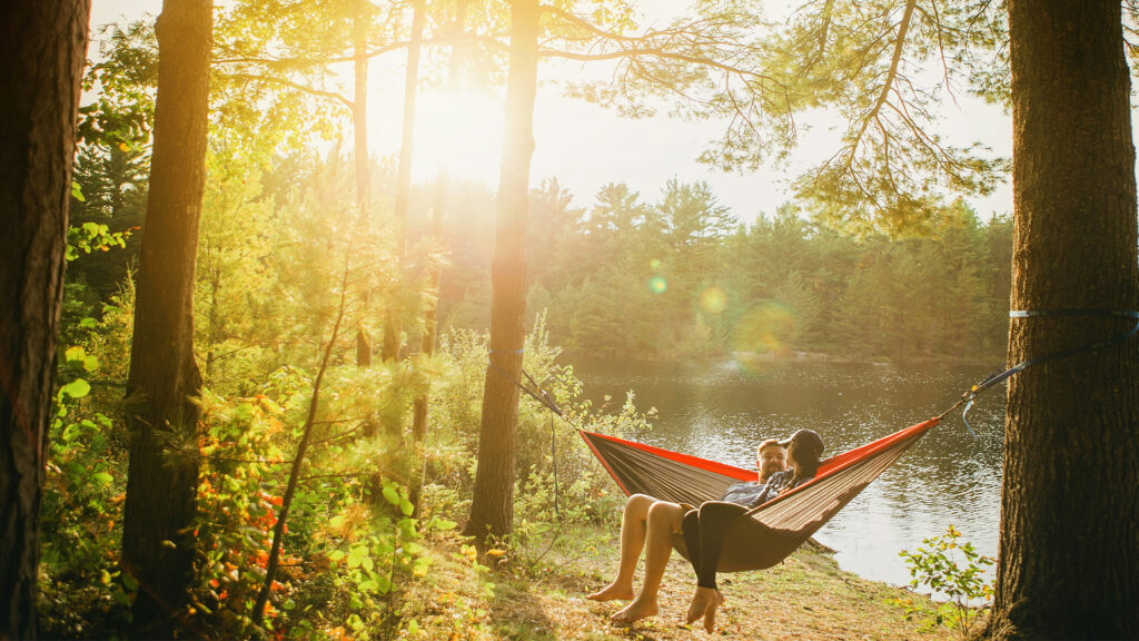 A couple on vacation sits in a hammock beside a forested lake.