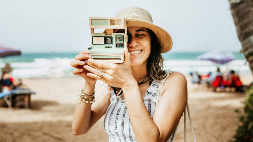 A woman holding a polaroid camera, taking a picture of the viewer.