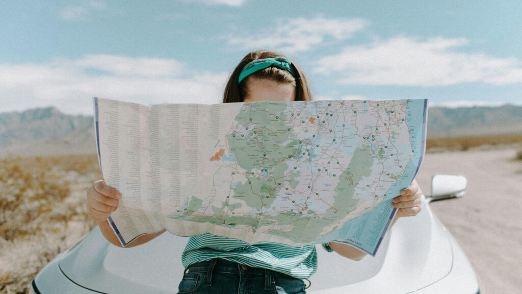 A woman holding a map, sitting on the hood of her car.