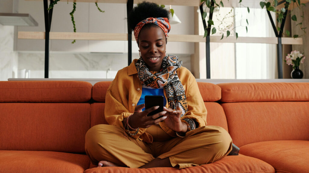 A woman sits on her couch, holding her cell phone and smiling.