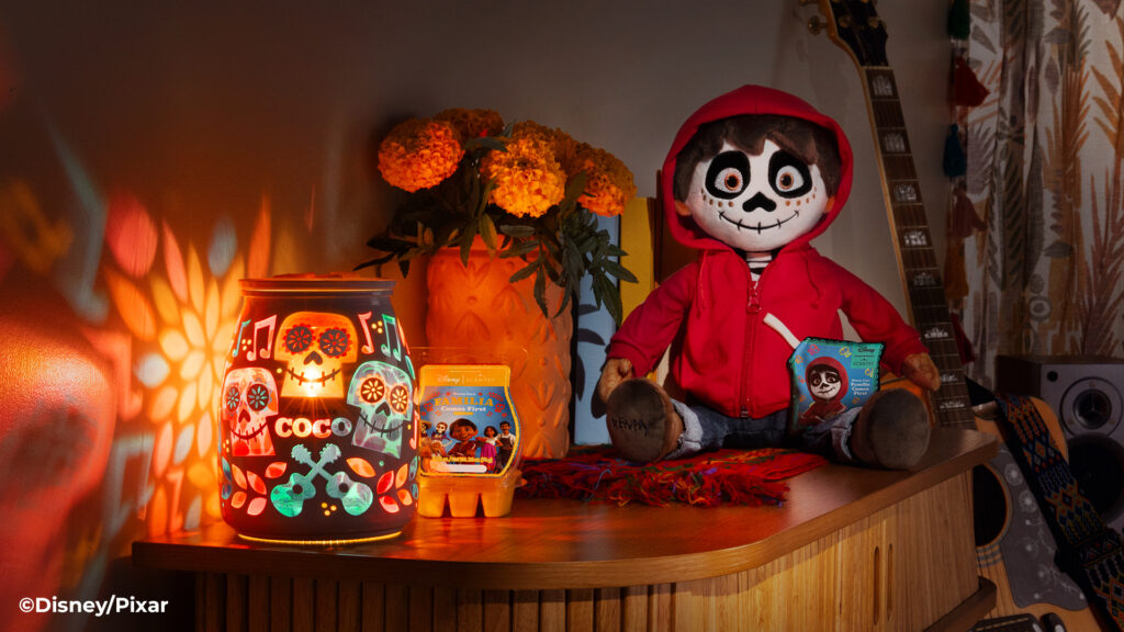 An artful display of the Disney Coco Scentsy Warmer, Disney Miguel Scentsy Buddy, and the Disney Coco: familia comes first Scentsy Bar, arranged beside marigold flowers. 