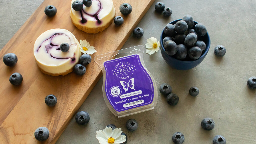 A Blueberry Cheesecake Scentsy Wax Bar sits on a table beside scattered blueberries and two small servings of blueberry cheesecake.