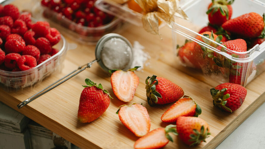 Sliced strawberries lay upon a cutting board surrounded by other different berries.