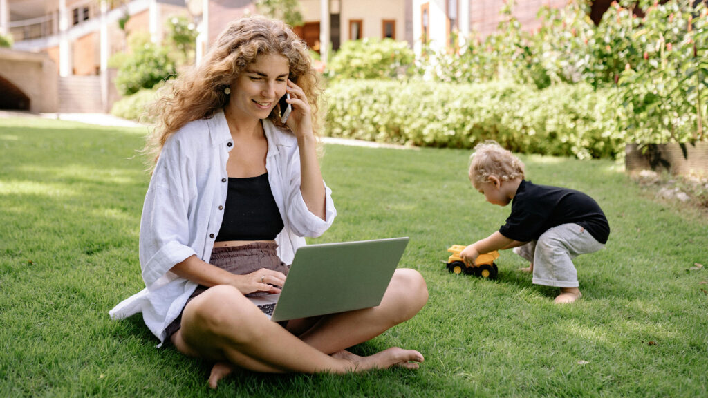 a woman works on a laptop and talks on the phone while sitting on the lawn in her yard, her toddler playing beside her.