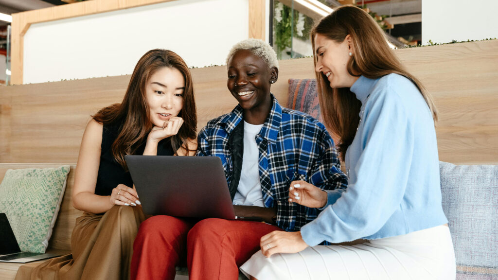 Three woman sit together around a laptop smiling at the screen.