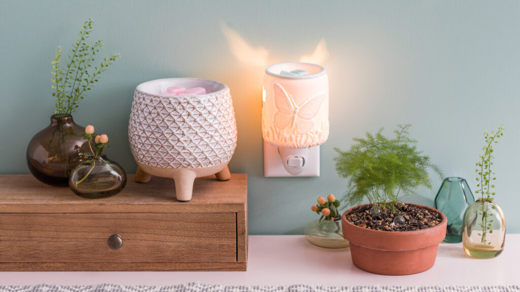 A Scentsy mini-warmer plugged into a wall outlet and another scentsy warmer next to natural plant decorations.