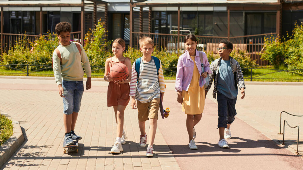 Five children with school bags, skateboards, and a basketball walk down the sidewalk together.