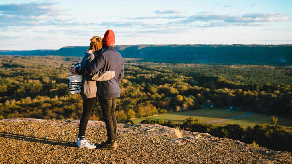 Two people standing on scenic overlook, holding each other as they gaze out over the countryside.