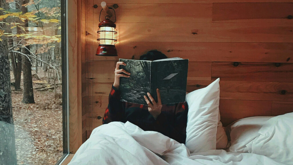 A person reading a book in their bed inside a rustic cabin.