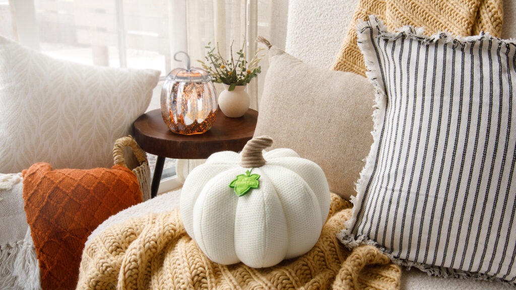 A cozy image of the Lumina Pumpkin Darling Decor pillow sitting on a soft couch with fuzzy blankets and soft cushions. 